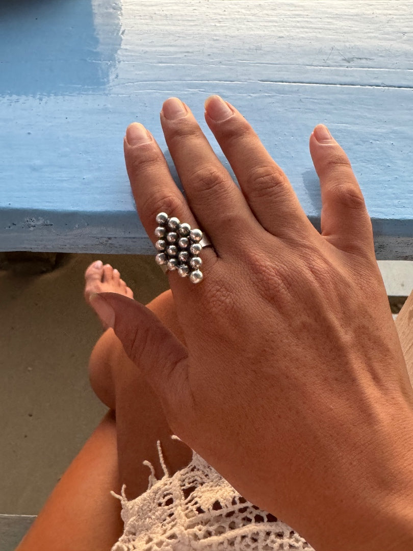 A close-up of a hand wearing a silver ring made of clustered small metallic beads, resting on a light blue wooden surface.