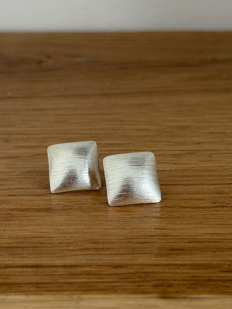 A pair of square, textured silver-toned stud earrings resting on a wooden surface.