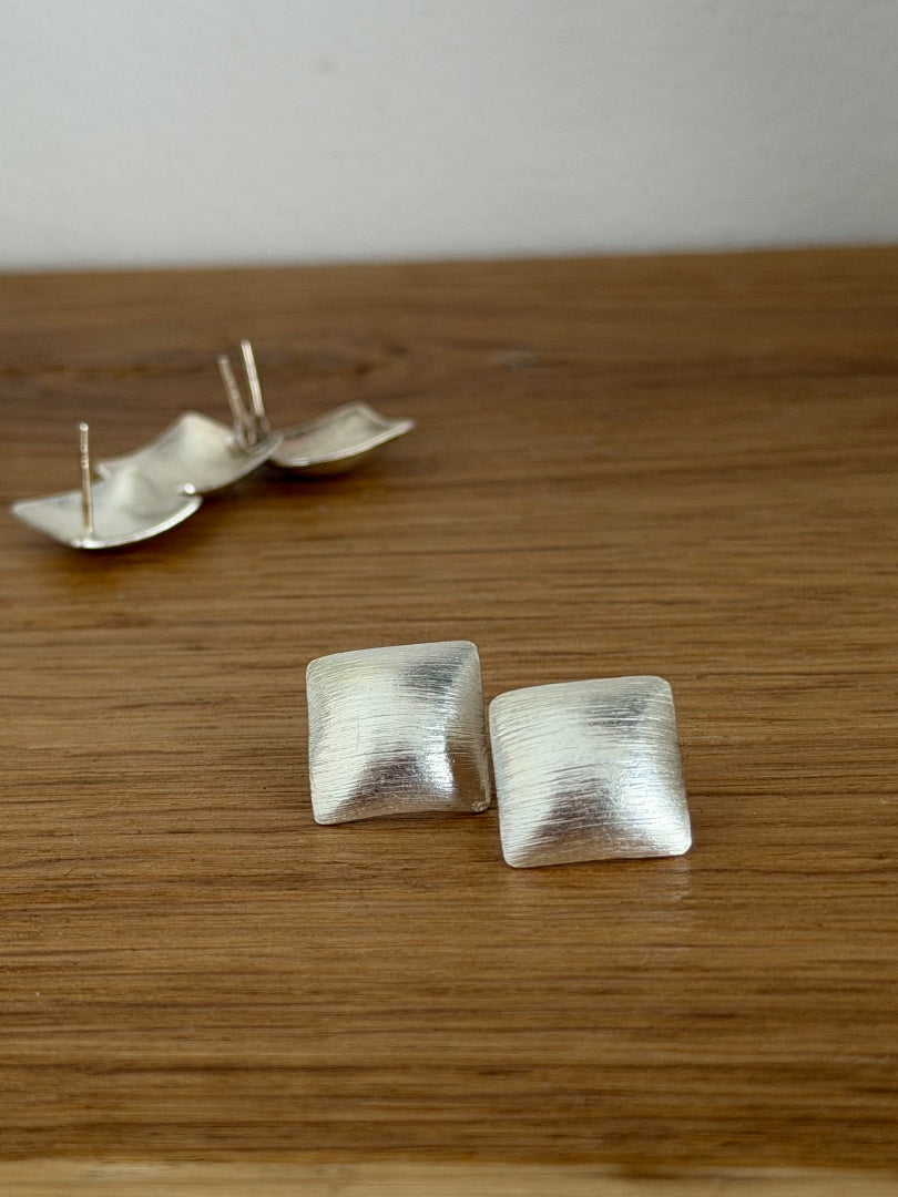A pair of textured silver square stud earrings resting on a wooden surface, with a pair of matching earrings shown in the blurred background.