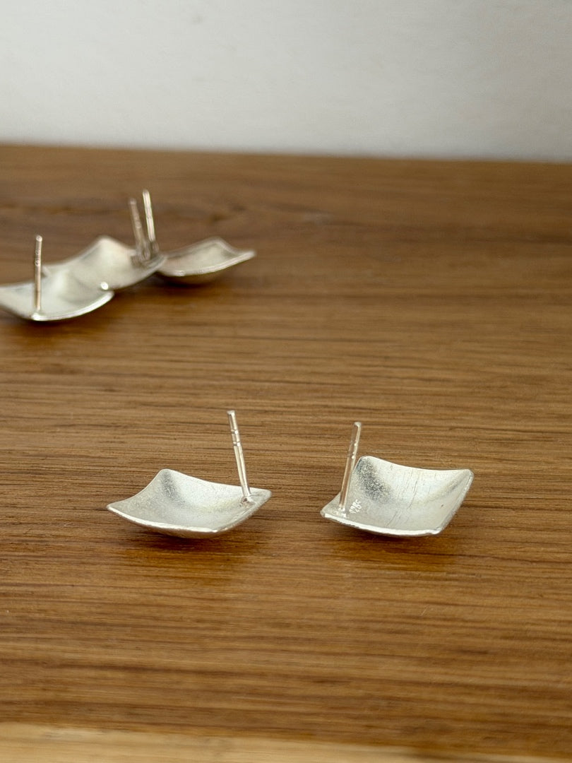 Three silver textured square stud earrings with flat posts, displayed on a wooden surface against a neutral light gray background.