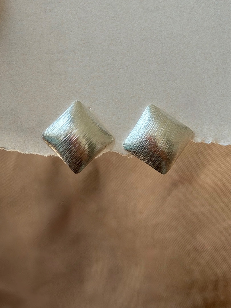 A pair of textured metallic square stud earrings with a brushed silver finish, resting on a torn paper background with a beige top half and a brown bottom half.