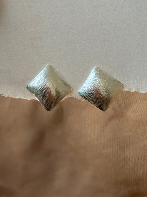 A pair of textured metallic square stud earrings with a brushed silver finish, resting on a torn paper background with a beige top half and a brown bottom half.