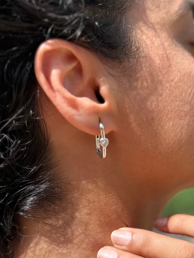 A close-up of a person's ear wearing a silver huggie hoop earring with a small clear heart-shaped crystal.