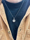 Close-up of a necklace with a pendant worn by a person in a blue shirt and beige jacket.