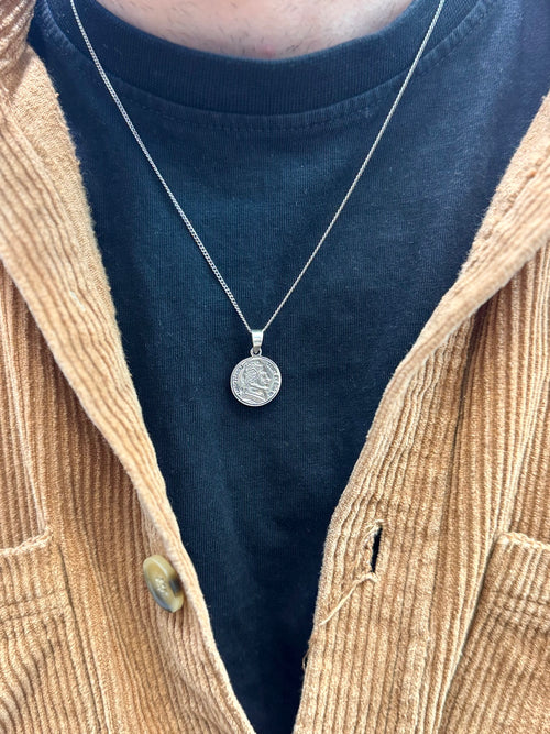 Close-up of a necklace with a pendant worn by a person in a blue shirt and beige jacket.