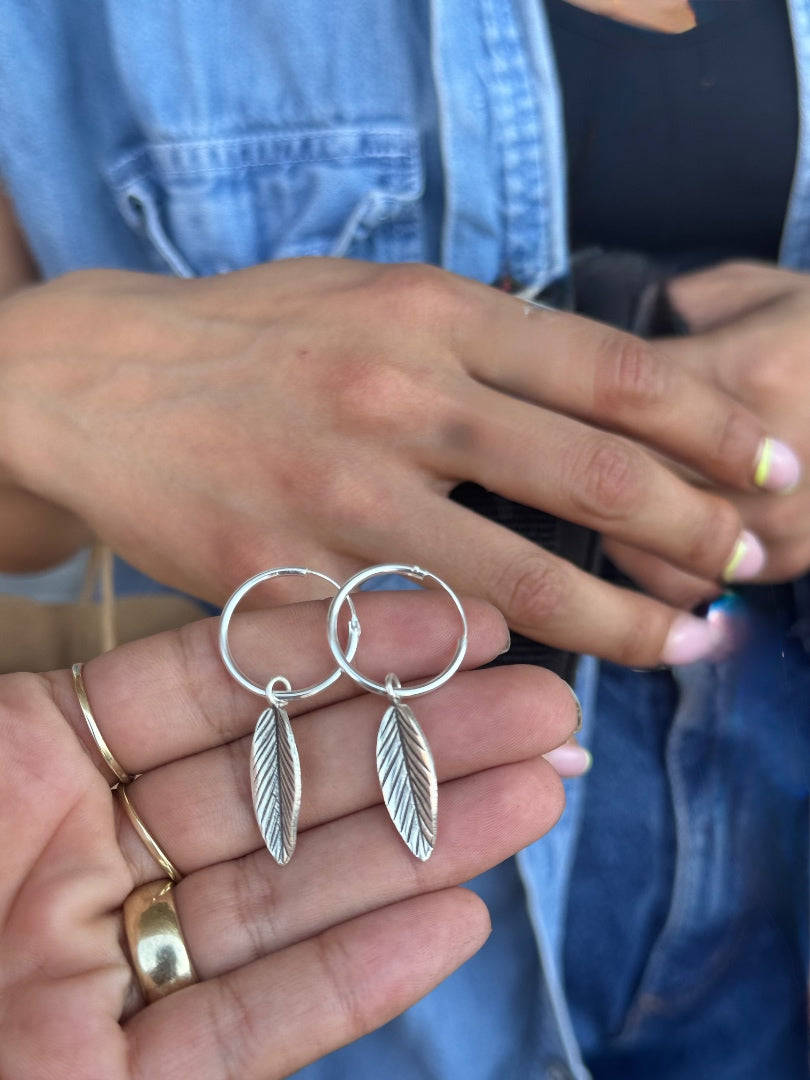 Silver hoop earrings with leaf designs held by a person wearing a denim jacket.