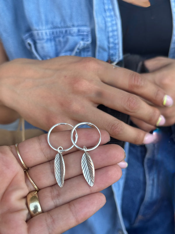 Silver hoop earrings with leaf designs held by a person wearing a denim jacket.