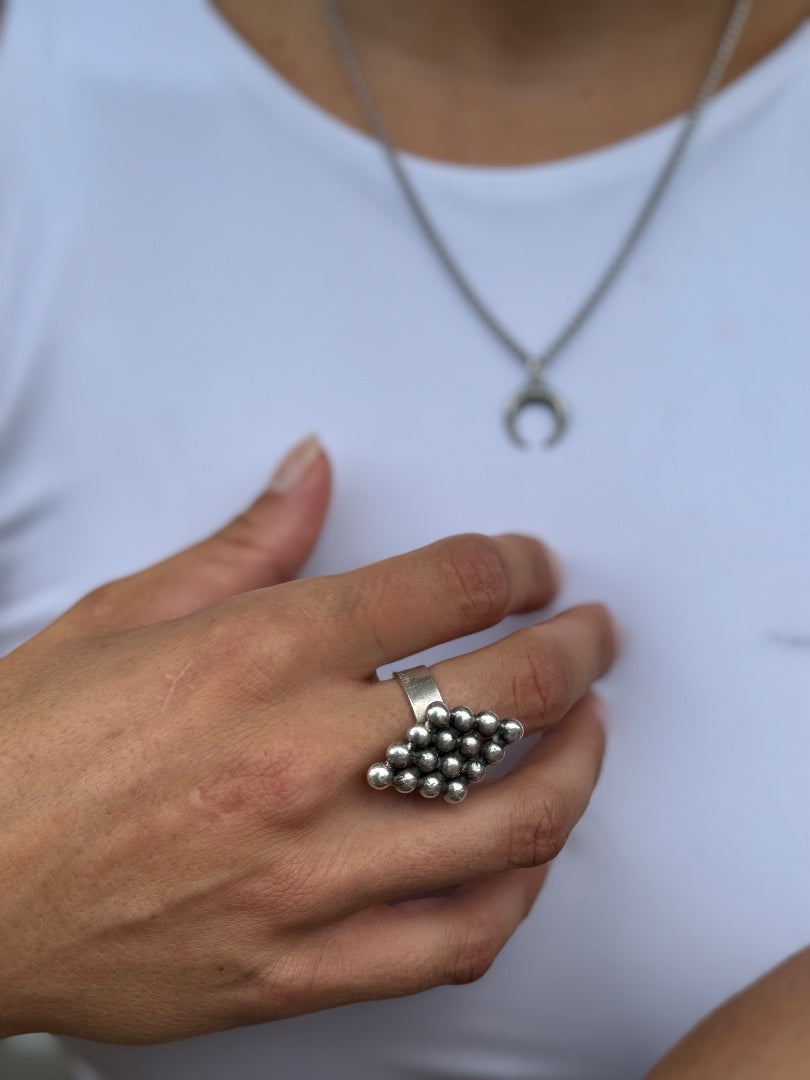 A close-up of a person's hand wearing a silver ring with a cluster of small metallic beads, against a white shirt background.