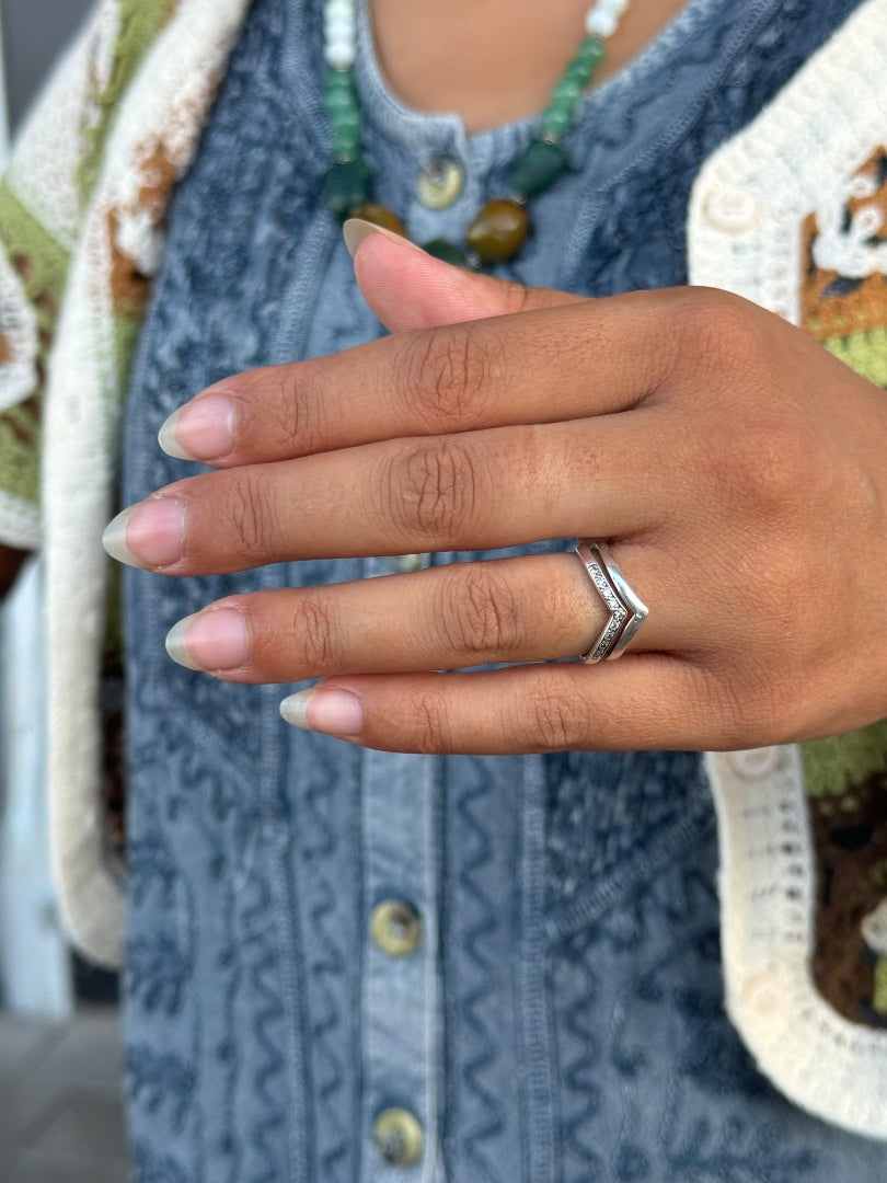 A close-up of a person's hand wearing a silver chevron ring, styled with a blue patterned top and a green beaded necklace.