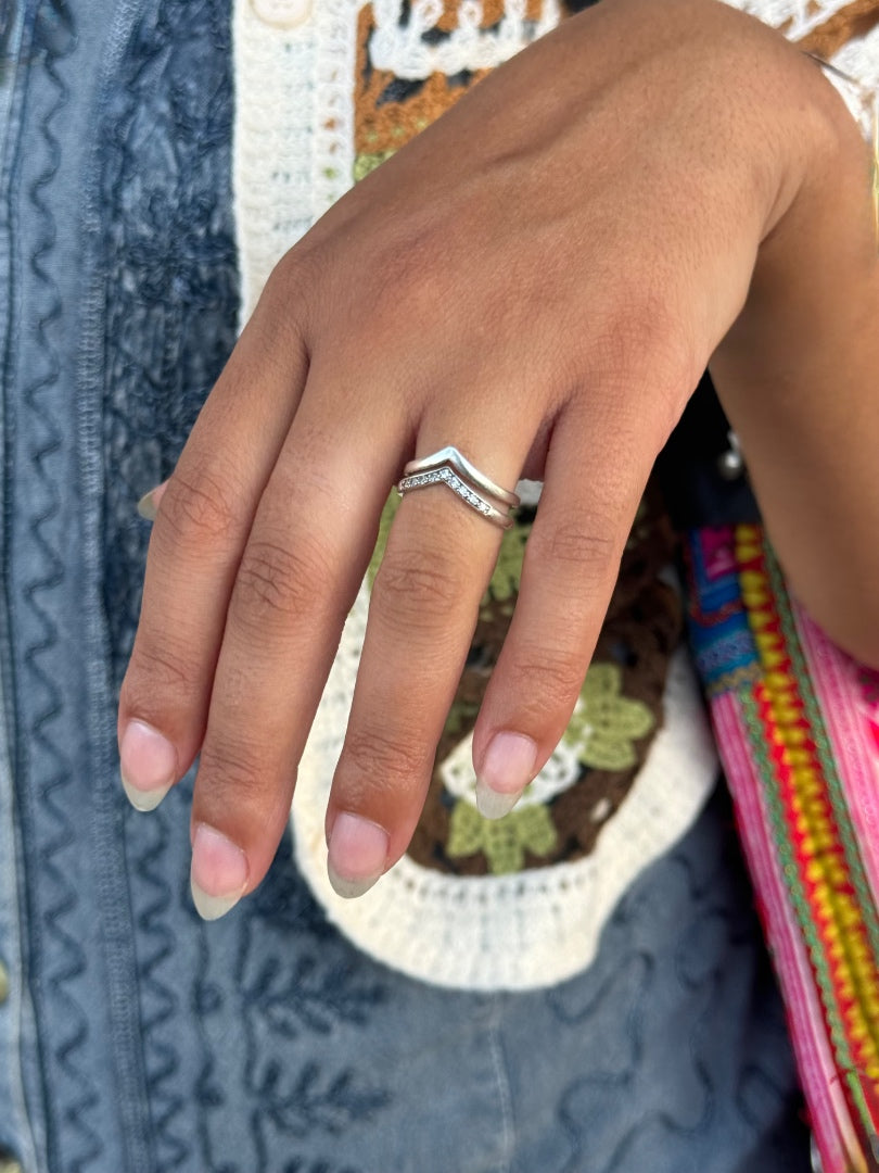 A close-up of a person's hand wearing a silver chevron ring with small clear stones, set against a background of patterned clothing.