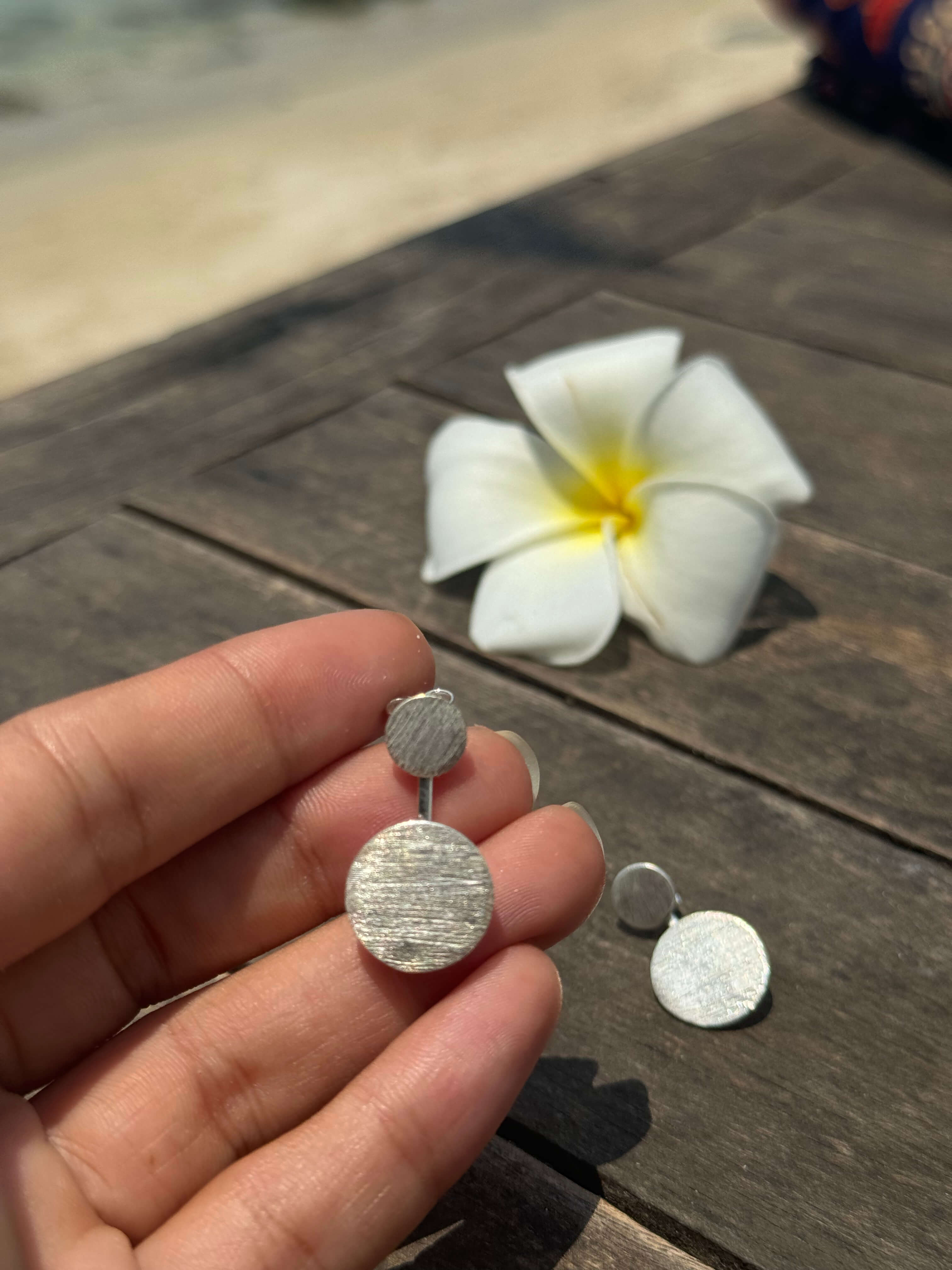 Silver earrings held in a hand with a white flower on a wooden surface