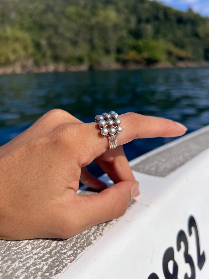 A silver ring with a cluster of small white pearls on a finger, shown outdoors on a boat with water and a green forested shoreline in the background.