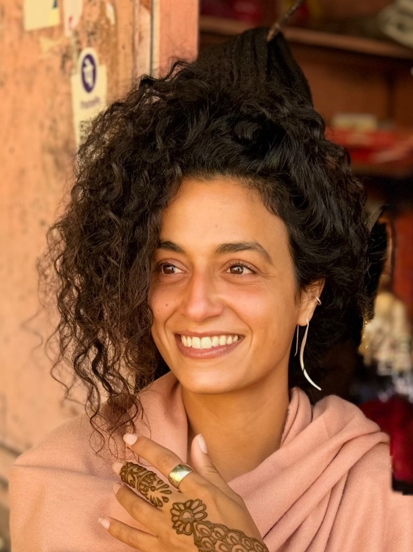 Woman with curly hair and henna on her hands, wearing a pink top, in a warm-toned indoor setting.