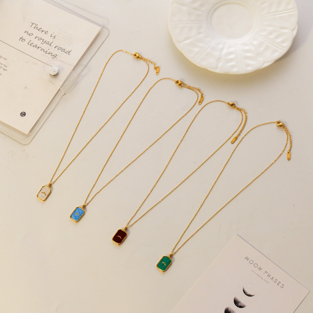 Four gold chain necklaces with rectangular pendant charms in blue, red, green, and gold, arranged on a light beige surface with decorative props including a white plate and cards.