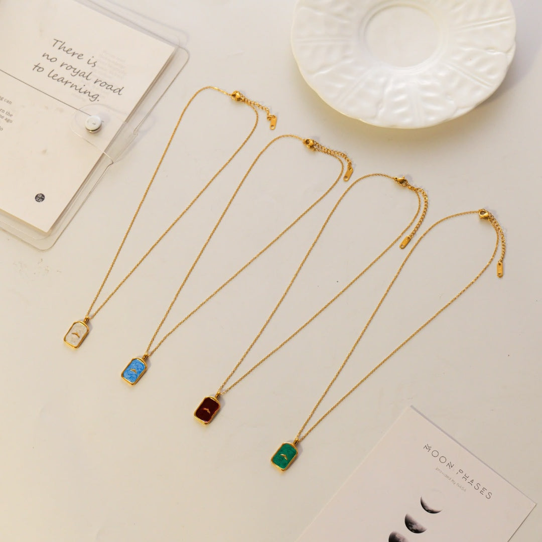 Four gold chain necklaces with rectangular pendant charms in blue, red, green, and gold, arranged on a light beige surface with decorative props including a white plate and cards.