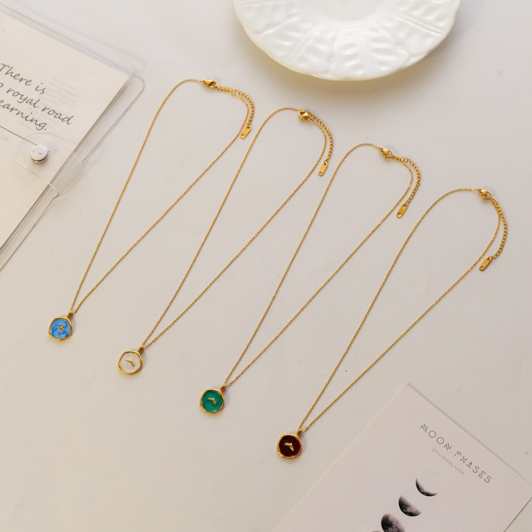 Four gold chain necklaces with round stone pendants in blue, gold, green, and dark red, arranged on a light background with decorative cards and a white dish.