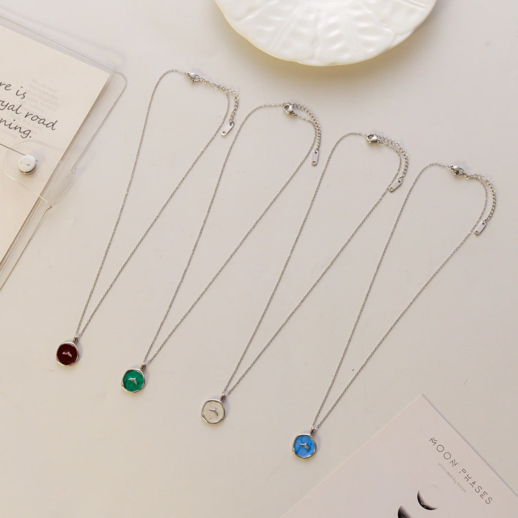 Four silver chain necklaces with round pendants in dark red, green, silver, and blue, arranged diagonally on a light beige surface with decorative props.