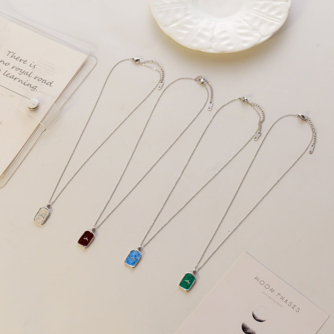 Four silver-toned pendant necklaces with rectangular charms in red, blue, and green, arranged on a light beige surface with decorative cards and a white dish.