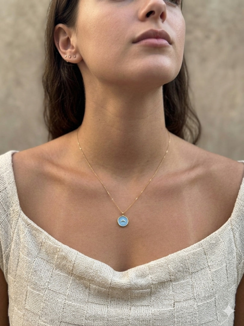A close-up of a woman wearing a delicate gold chain necklace with a small round light blue pendant, paired with a textured off-white top against a neutral beige background.